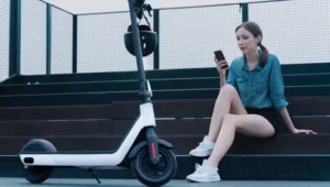 Eine junge Dame sitzt auf einer Treppe und sieht auf ihr Handy. Neben ihr parkt ihr E-Scooter.