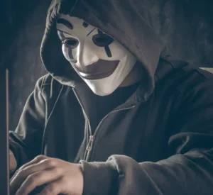 Eine Person mit Clownsmaske sitzt vor einem Computer. Es scheint ein betrügerischer Hacker zu sein.